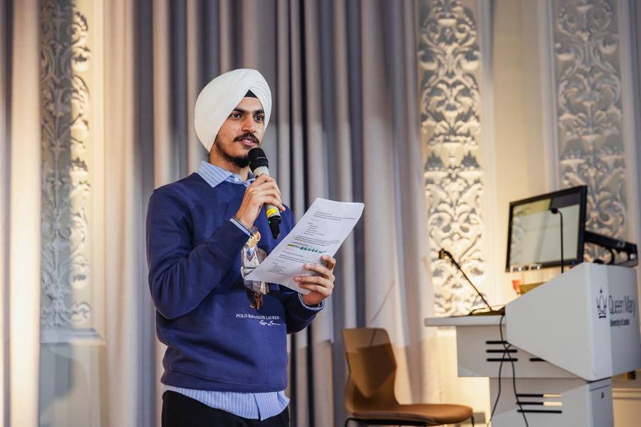 Eshwinder speaking with a microphone and notes at a QMUL event podium